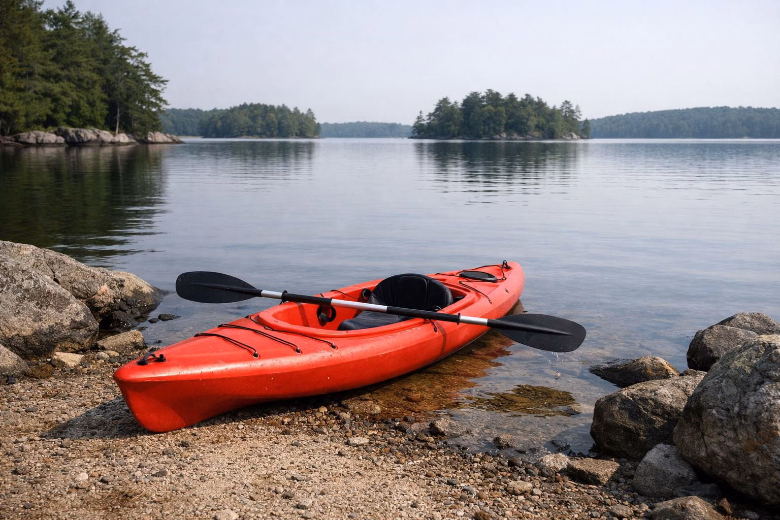 An empty red kayak with a paddle pulled up on a rocky lake shore, wooded islands beyond